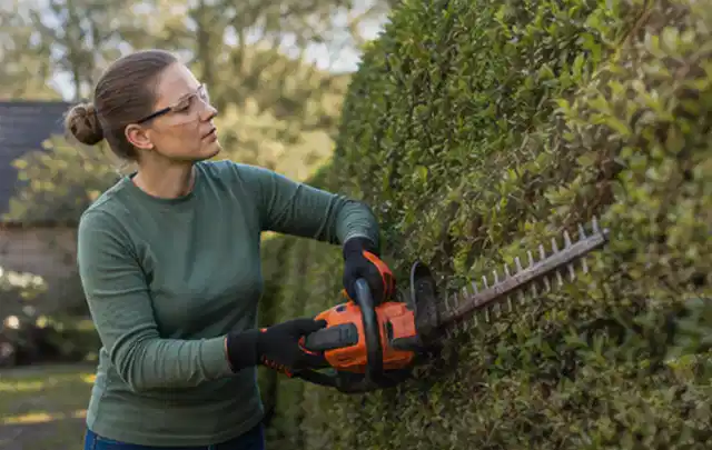 Person schneidet eine dichte Gartenhecke mit Schutzbrille und sicherem Stand; Thema Akku Gartengeräte, Schwerpunkt Dimensionierung; Umsetzung.