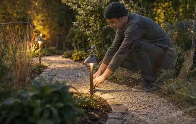 Person richtet eine niedrige warm leuchtende Gartenlampe am Kiesweg aus; Thema Aufstellpools, Schwerpunkt Anwendung; Kontrolle.