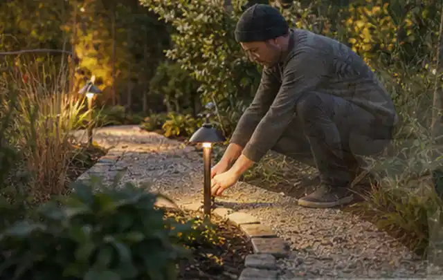 Person richtet eine niedrige warm leuchtende Gartenlampe am Kiesweg aus; Thema Aufstellpools, Schwerpunkt Checkliste; Kontrolle.