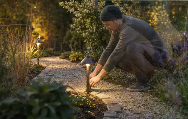 Person richtet eine niedrige warm leuchtende Gartenlampe am Kiesweg aus; Thema Aufstellpools, Schwerpunkt Ersatzteile; Ausgangslage.