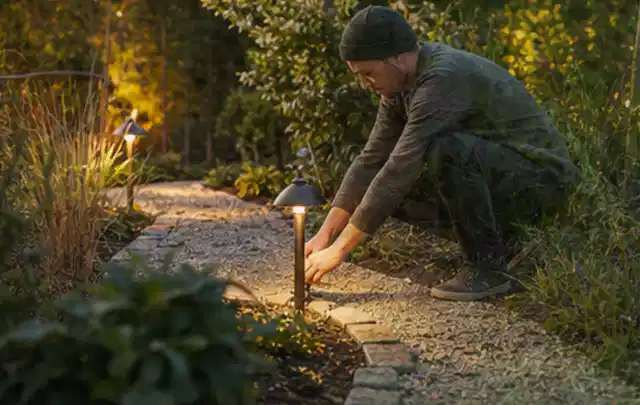 Person richtet eine niedrige warm leuchtende Gartenlampe am Kiesweg aus; Thema Aufstellpools, Schwerpunkt Kaufberatung; Ergebnis.