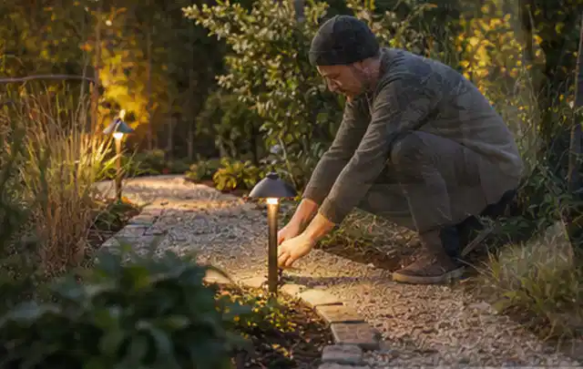 Person richtet eine niedrige warm leuchtende Gartenlampe am Kiesweg aus; Thema Aufstellpools, Schwerpunkt Reinigung; Praxisalltag.