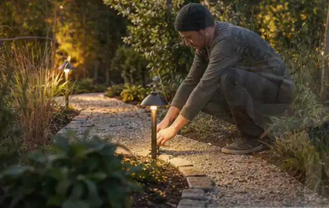 Person richtet eine niedrige warm leuchtende Gartenlampe am Kiesweg aus; Thema Aufstellpools, Schwerpunkt Sicherheit; Umsetzung.