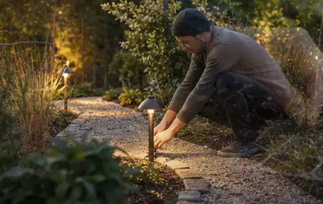 Person richtet eine niedrige warm leuchtende Gartenlampe am Kiesweg aus; Thema Aufstellpools, Schwerpunkt Systemvergleich; Praxisalltag.