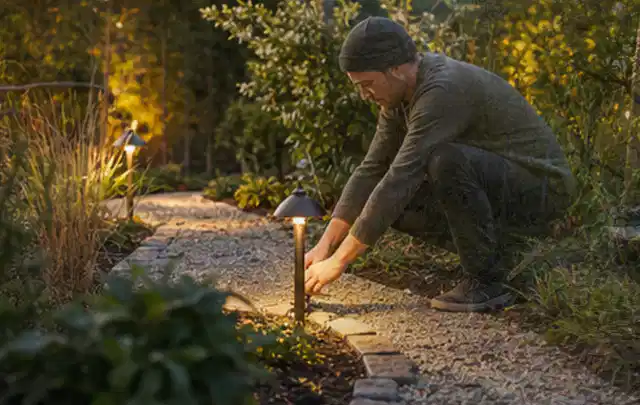 Person richtet eine niedrige warm leuchtende Gartenlampe am Kiesweg aus; Thema Aufstellpools, Schwerpunkt Testkriterien; Praxisalltag.