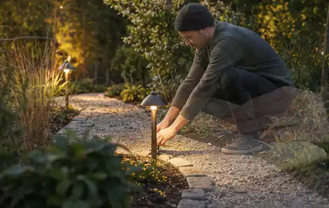 Person richtet eine niedrige warm leuchtende Gartenlampe am Kiesweg aus; Thema Balkon, Schwerpunkt Gärten mit kleiner Fläche; Praxisalltag.
