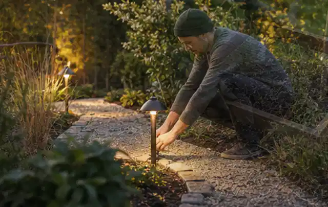 Person richtet eine niedrige warm leuchtende Gartenlampe am Kiesweg aus; Thema Balkon, Schwerpunkt Inspiration; Umsetzung.