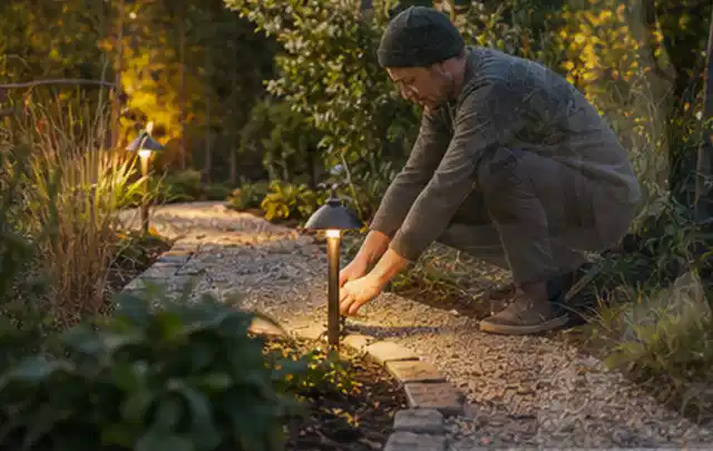 Person richtet eine niedrige warm leuchtende Gartenlampe am Kiesweg aus; Thema Balkon, Schwerpunkt Materialwahl; Ergebnis.