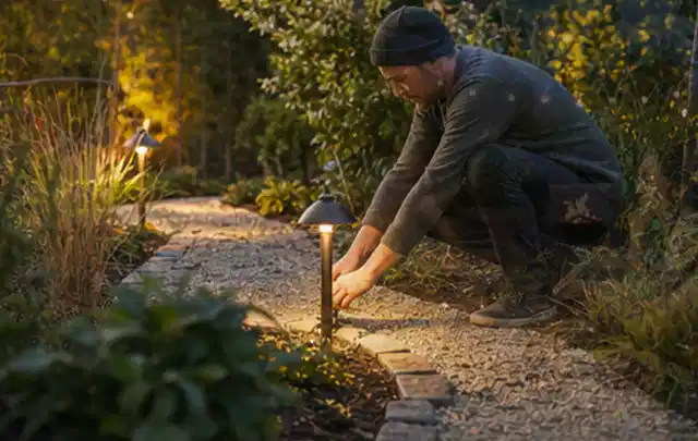 Person richtet eine niedrige warm leuchtende Gartenlampe am Kiesweg aus; Thema Balkon, Schwerpunkt Naturgarten; Kontrolle.