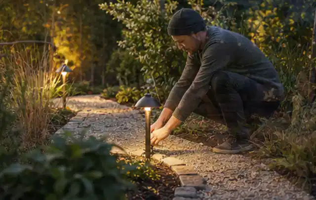 Person richtet eine niedrige warm leuchtende Gartenlampe am Kiesweg aus; Thema Balkon, Schwerpunkt Praxisbeispiele; Umsetzung.