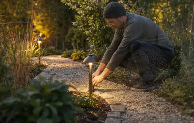 Person richtet eine niedrige warm leuchtende Gartenlampe am Kiesweg aus; Thema Bambus Sichtschutz, Schwerpunkt DIY; Praxisalltag.