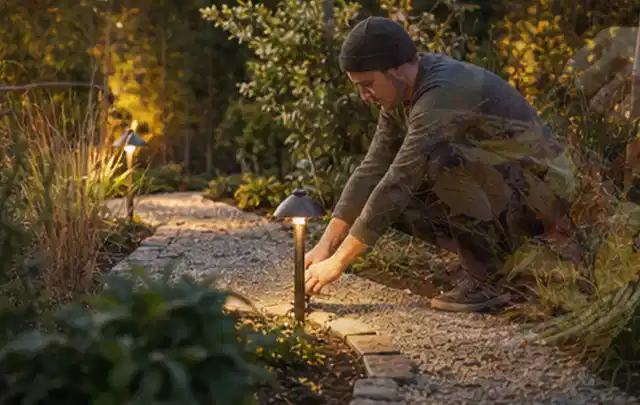 Person richtet eine niedrige warm leuchtende Gartenlampe am Kiesweg aus; Thema Bambus Sichtschutz, Schwerpunkt Gestaltung; Umsetzung.