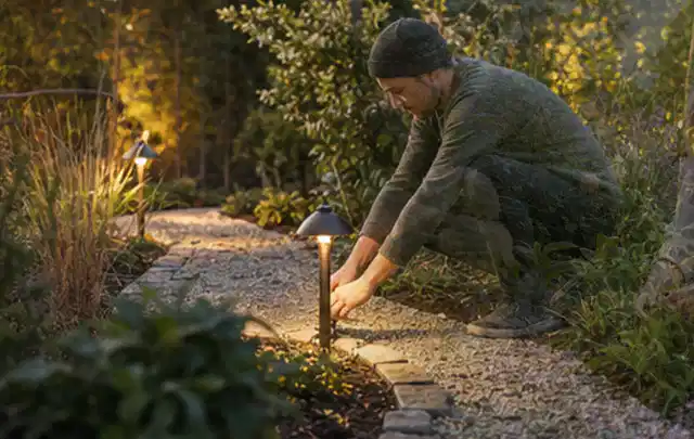 Person richtet eine niedrige warm leuchtende Gartenlampe am Kiesweg aus; Thema Bambus Sichtschutz, Schwerpunkt Materialwahl; Umsetzung.