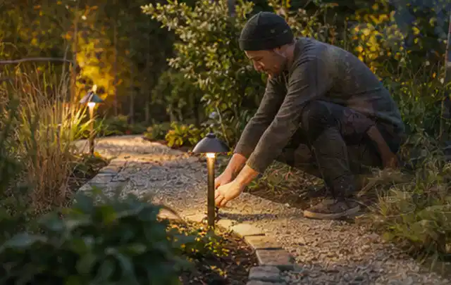 Person richtet eine niedrige warm leuchtende Gartenlampe am Kiesweg aus; Thema Bambus Sichtschutz, Schwerpunkt Pflege; Umsetzung.