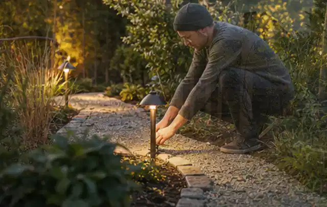 Person richtet eine niedrige warm leuchtende Gartenlampe am Kiesweg aus; Thema Bambus Sichtschutz, Schwerpunkt Standort; Umsetzung.