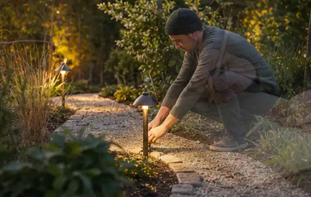 Person richtet eine niedrige warm leuchtende Gartenlampe am Kiesweg aus; Thema Bambus Sichtschutz, Schwerpunkt Winterschutz; Kontrolle.