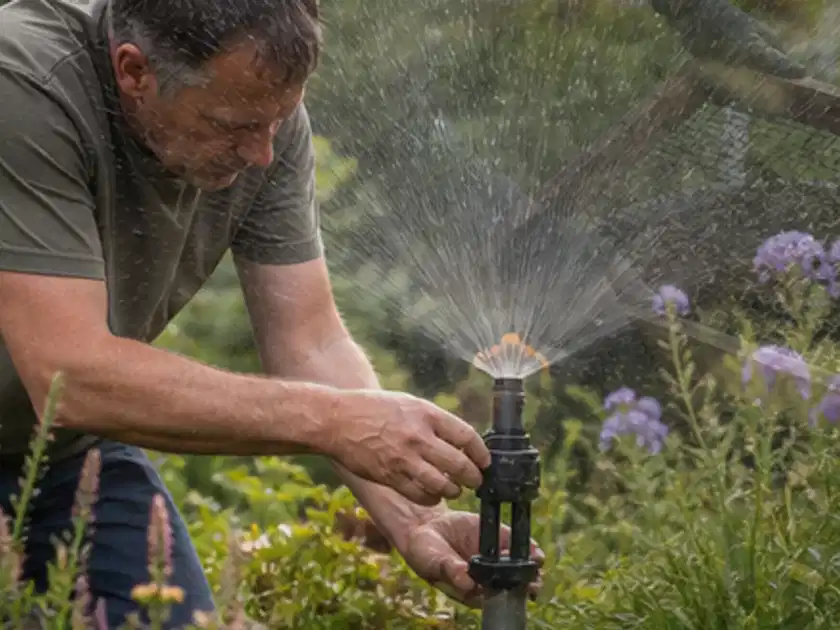 Person richtet einen laufenden Gartensprenger zwischen Stauden präzise aus; Thema Bewässerungscomputer, Schwerpunkt Leistung; Titelbild.
