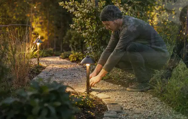 Person richtet eine niedrige warm leuchtende Gartenlampe am Kiesweg aus; Thema Bosch Indego, Schwerpunkt Kosten; Praxisalltag.