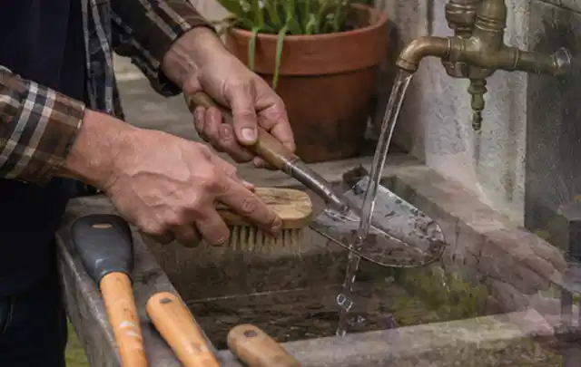 Hände reinigen gebrauchte Gartengeräte mit Bürste und Wasser am Außenbecken; Thema Bosch Indego, Schwerpunkt Reinigung; Kontrolle.