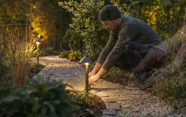 Person richtet eine niedrige warm leuchtende Gartenlampe am Kiesweg aus; Thema Bosch Indego, Schwerpunkt Reparatur; Ergebnis.