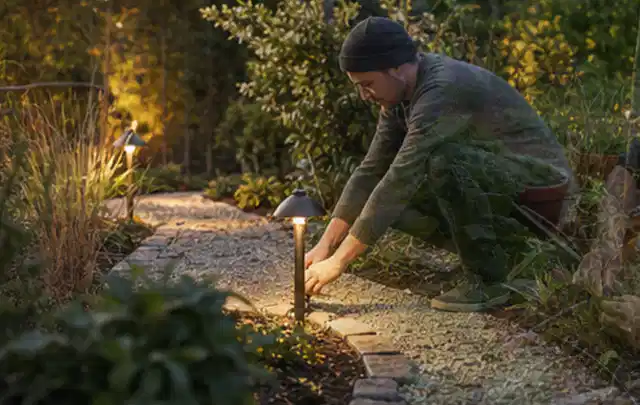Person richtet eine niedrige warm leuchtende Gartenlampe am Kiesweg aus; Thema Bosch Indego, Schwerpunkt Winterlager; Ergebnis.