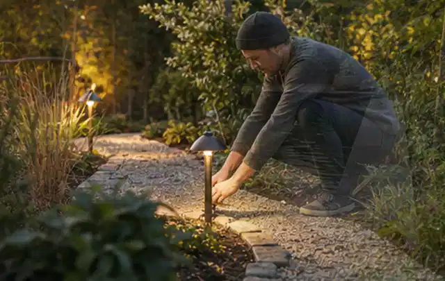 Person richtet eine niedrige warm leuchtende Gartenlampe am Kiesweg aus; Thema Carports, Schwerpunkt Fehlerhilfe; Ergebnis.