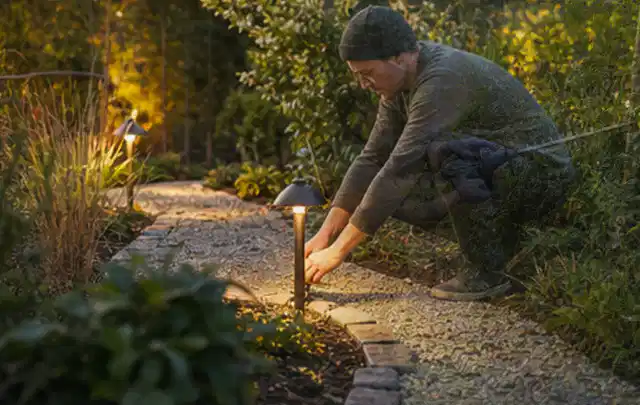 Person richtet eine niedrige warm leuchtende Gartenlampe am Kiesweg aus; Thema Carports, Schwerpunkt Gestaltung; Praxisalltag.