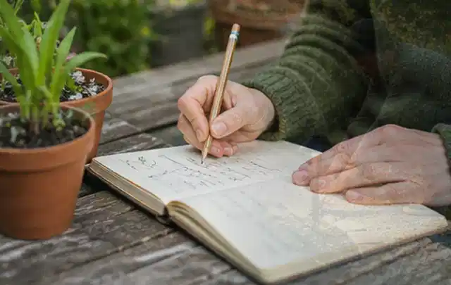 Person dokumentiert Pflanzenbeobachtungen in einem Gartentagebuch am Holztisch; Thema DIY, Schwerpunkt Ratgeber; Umsetzung.