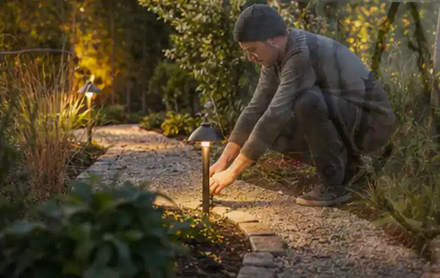 Person richtet eine niedrige warm leuchtende Gartenlampe am Kiesweg aus; Thema Dreame A1, Schwerpunkt Checkliste; Praxisalltag.