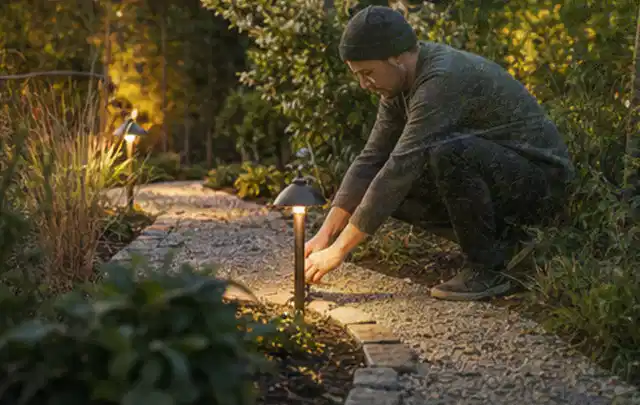 Person richtet eine niedrige warm leuchtende Gartenlampe am Kiesweg aus; Thema Dreame A1, Schwerpunkt Dimensionierung; Umsetzung.