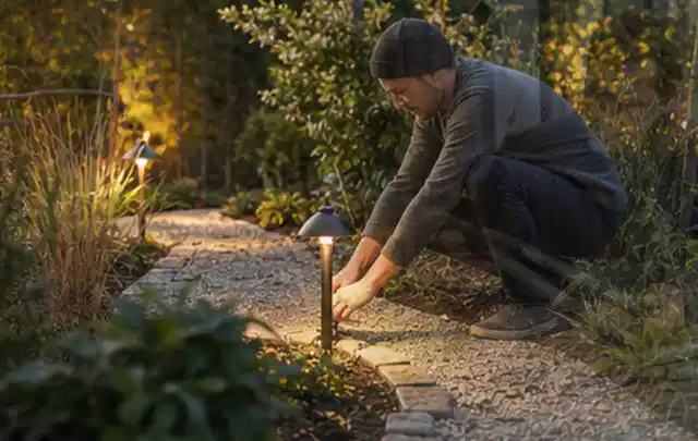 Person richtet eine niedrige warm leuchtende Gartenlampe am Kiesweg aus; Thema Dreame A1, Schwerpunkt Gärten mit großer Fläche; Praxisalltag.