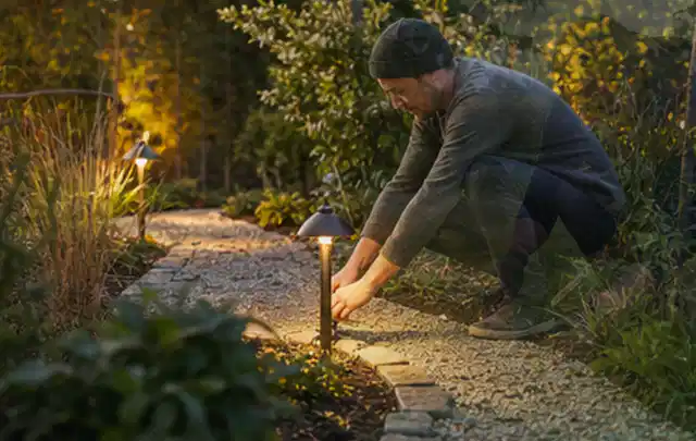 Person richtet eine niedrige warm leuchtende Gartenlampe am Kiesweg aus; Thema Dreame A1, Schwerpunkt Gärten mit kleiner Fläche; Praxisalltag.