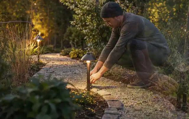 Person richtet eine niedrige warm leuchtende Gartenlampe am Kiesweg aus; Thema Dreame A1, Schwerpunkt Nachhaltigkeit; Ausgangslage.