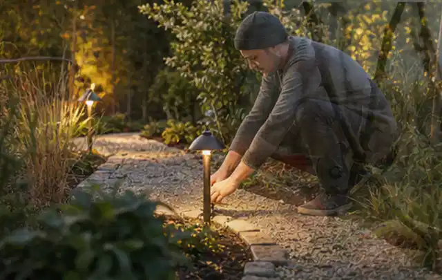 Person richtet eine niedrige warm leuchtende Gartenlampe am Kiesweg aus; Thema Dreame A1, Schwerpunkt Testkriterien; Umsetzung.