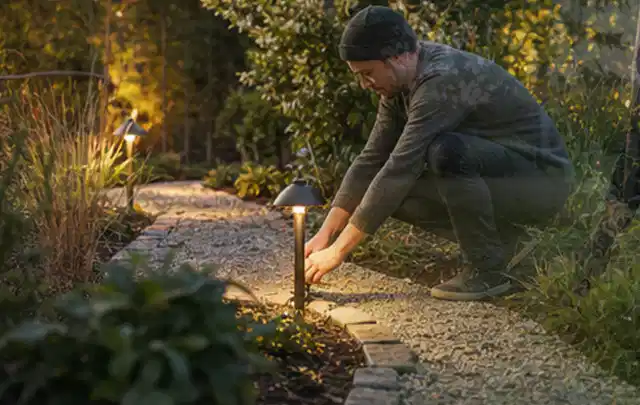 Person richtet eine niedrige warm leuchtende Gartenlampe am Kiesweg aus; Thema Ecovacs Goat, Schwerpunkt Anwendung; Umsetzung.