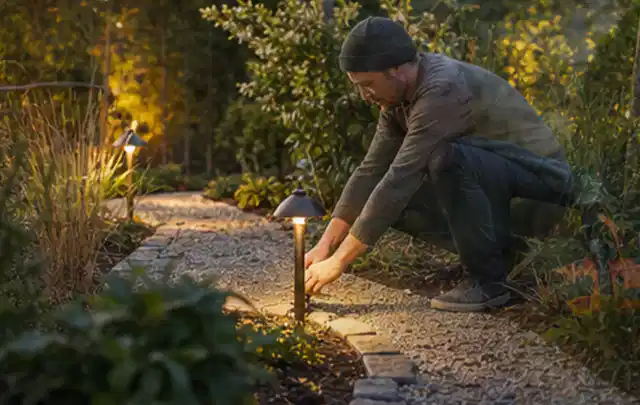 Person richtet eine niedrige warm leuchtende Gartenlampe am Kiesweg aus; Thema Ecovacs Goat, Schwerpunkt Bestenliste; Kontrolle.