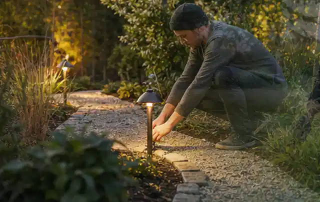 Person richtet eine niedrige warm leuchtende Gartenlampe am Kiesweg aus; Thema Ecovacs Goat, Schwerpunkt Checkliste; Ergebnis.