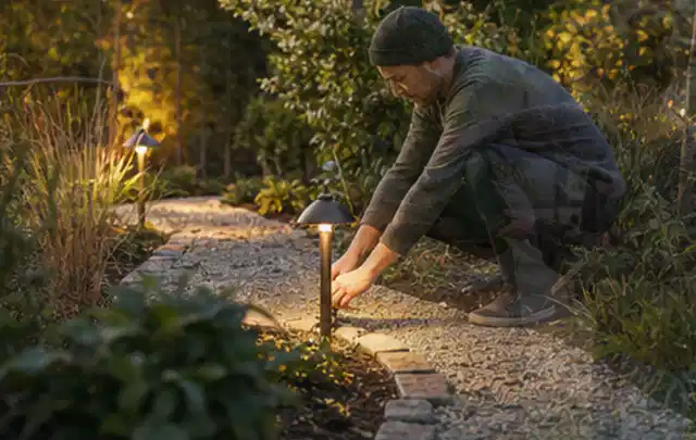 Person richtet eine niedrige warm leuchtende Gartenlampe am Kiesweg aus; Thema Ecovacs Goat, Schwerpunkt Gärten mit kleiner Fläche; Praxisalltag.
