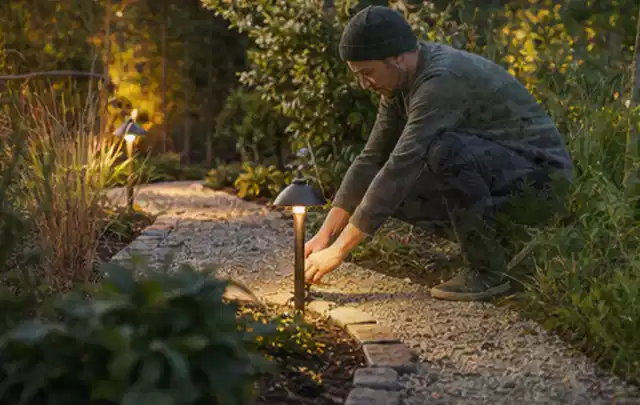 Person richtet eine niedrige warm leuchtende Gartenlampe am Kiesweg aus; Thema Ecovacs Goat, Schwerpunkt Leistung; Ergebnis.