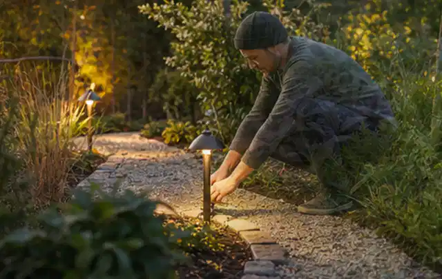 Person richtet eine niedrige warm leuchtende Gartenlampe am Kiesweg aus; Thema Ecovacs Goat, Schwerpunkt Nachhaltigkeit; Praxisalltag.