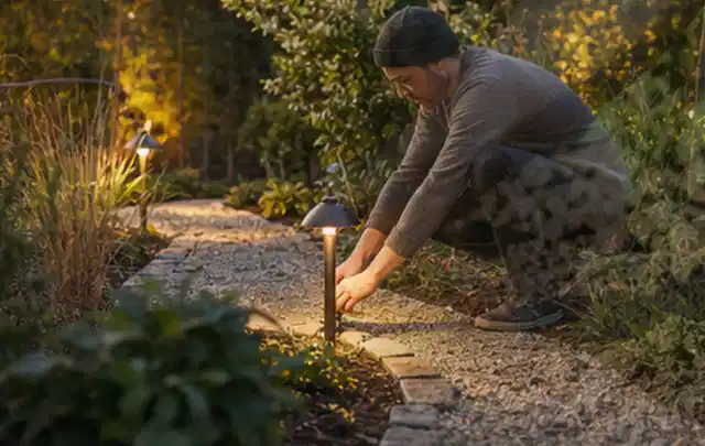 Person richtet eine niedrige warm leuchtende Gartenlampe am Kiesweg aus; Thema Ecovacs Goat, Schwerpunkt Reinigung; Kontrolle.