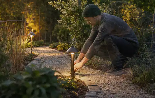 Person richtet eine niedrige warm leuchtende Gartenlampe am Kiesweg aus; Thema Ecovacs Goat, Schwerpunkt Systemvergleich; Ausgangslage.