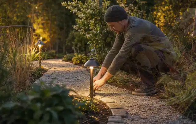 Person richtet eine niedrige warm leuchtende Gartenlampe am Kiesweg aus; Thema Einbaupools, Schwerpunkt Anwendung; Ergebnis.