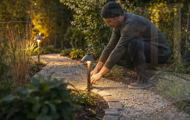 Person richtet eine niedrige warm leuchtende Gartenlampe am Kiesweg aus; Thema Einbaupools, Schwerpunkt Checkliste; Kontrolle.