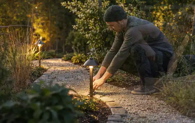 Person richtet eine niedrige warm leuchtende Gartenlampe am Kiesweg aus; Thema Einbaupools, Schwerpunkt Dimensionierung; Ausgangslage.