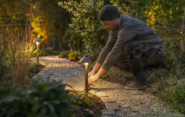Person richtet eine niedrige warm leuchtende Gartenlampe am Kiesweg aus; Thema Einbaupools, Schwerpunkt Fehlerhilfe; Praxisalltag.