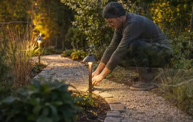 Person richtet eine niedrige warm leuchtende Gartenlampe am Kiesweg aus; Thema Einbaupools, Schwerpunkt Gärten mit kleiner Fläche; Praxisalltag.