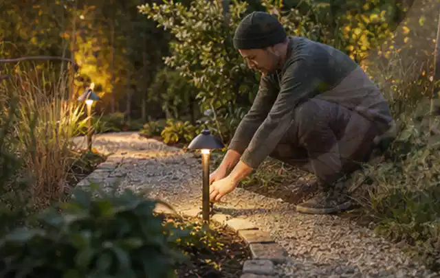 Person richtet eine niedrige warm leuchtende Gartenlampe am Kiesweg aus; Thema Einbaupools, Schwerpunkt Kaufberatung; Ausgangslage.