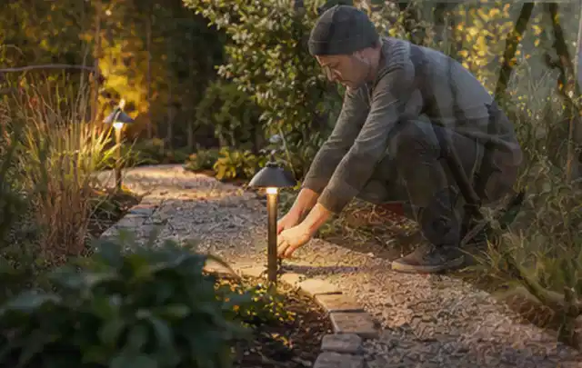 Person richtet eine niedrige warm leuchtende Gartenlampe am Kiesweg aus; Thema Einbaupools, Schwerpunkt Kosten; Praxisalltag.