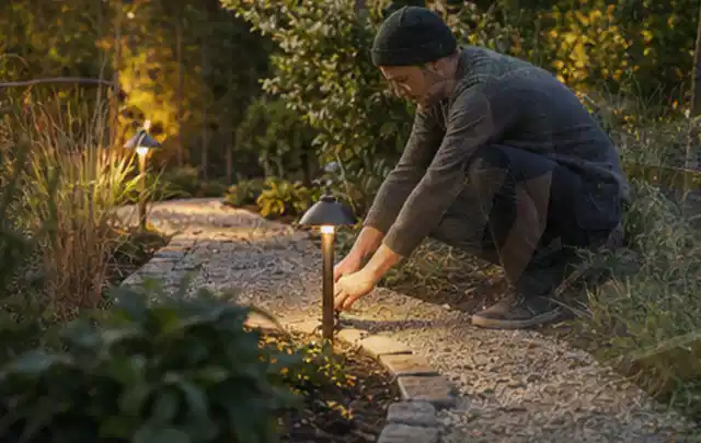 Person richtet eine niedrige warm leuchtende Gartenlampe am Kiesweg aus; Thema Einbaupools, Schwerpunkt Nachhaltigkeit; Kontrolle.